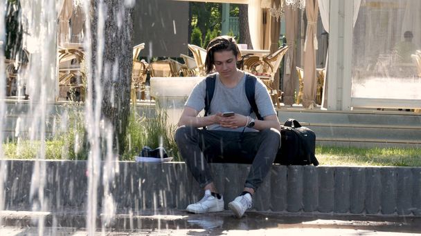 A young handsome guy (man) sits next to a fountain, writes a message on a mobile phone. Concept: young teenagers, lifestyle, schoolchild, student, park, summer. - Фото, зображення