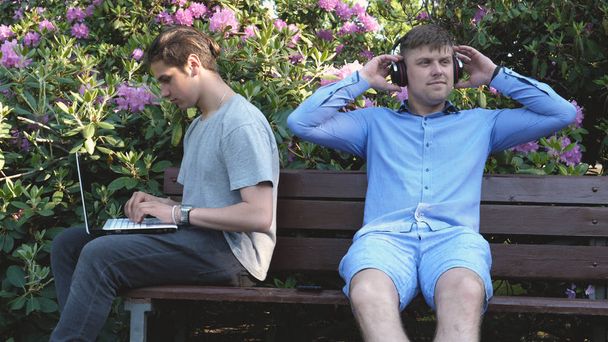 zwei schöne junge Leute Teenager, lesen ein Buch, hören Musik in einem Mobiltelefon, sitzen auf einer Bank, Hintergrund von Blumen und Wucherungen. Konzept: Studium, Studenten, Frisuren, Lebensstil. - Foto, Bild