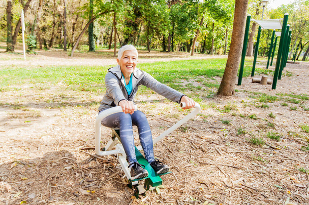 ältere Frau in Sportkleidung trainiert im Outdoor-Fitnesspark mit Rudergerät, gesunde Lebensweise Senioren. - Foto, Bild