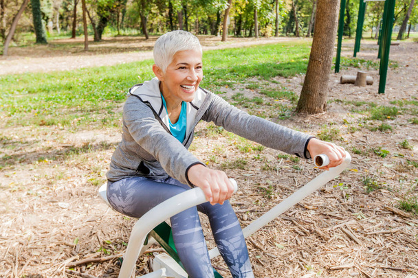 Femme âgée en vêtements de sport faisant de l'exercice au parc de remise en forme extérieur avec rameur, mode de vie sain Personnes âgées
. - Photo, image