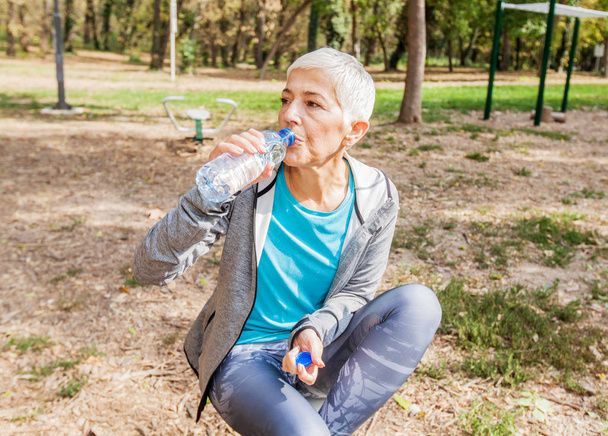 Femme âgée en bonne santé se détendre et boire de l'eau de la bouteille après l'entraînement au parc de remise en forme extérieur
 - Photo, image