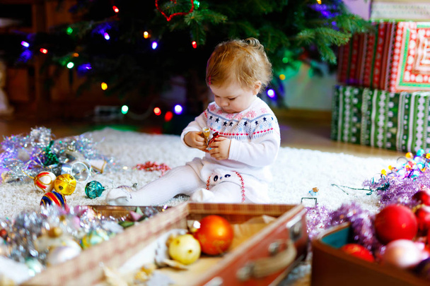 Αξιολάτρευτο κοριτσάκι κρατώντας πολύχρωμα vintage xmas παιχνίδι και η μπάλα στα χέρια χαριτωμένο. Μικρό παιδί σε γιορτινά ρούχα διακόσμηση χριστουγεννιάτικο δέντρο με την οικογένεια. Πρώτος εορτασμός του παραδοσιακό γλέντι - Φωτογραφία, εικόνα
