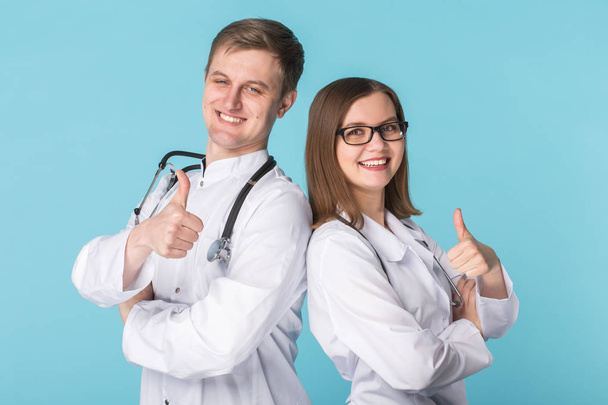 Два лучших умных профессиональных улыбающихся врача работников в белых халатах, показывающих большие пальцы, стоящие на синем фоне
 - Фото, изображение