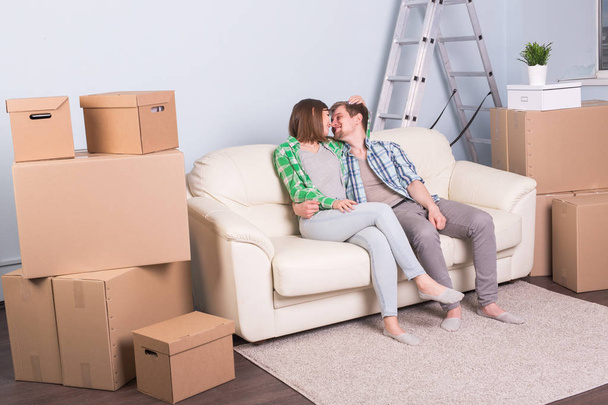 New home, relocation and moving concept - young couple sitting and kissing on the couch surrounded with boxes - Photo, Image