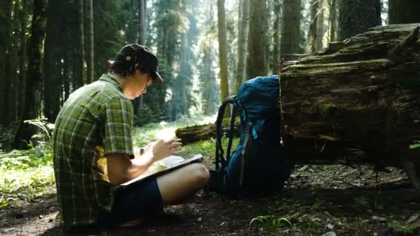 Tourist mit Rucksack sitzt im Wald und telefoniert, überprüft die Route per GPS und Karte. Mann erschrak über Bodenwellen, erschrak und duckte sich - Filmmaterial, Video