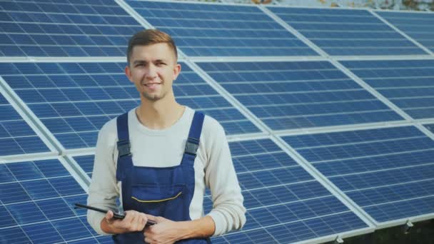Portrait of a young worker in workwear, smiling and looking at the camera. Against the background of solar panels - Záběry, video