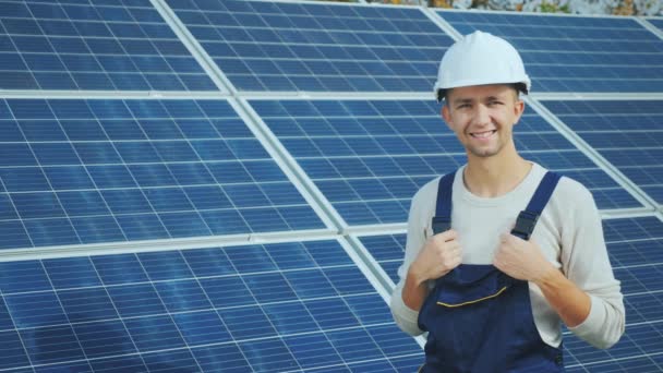 Portrait of a worker in a helmet, looking at the camera, stands against the background of a row of solar panels. alternative energy - Materiał filmowy, wideo