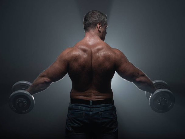 Powerful muscular man trains with dumbbells on black background in smoke - Fotó, kép