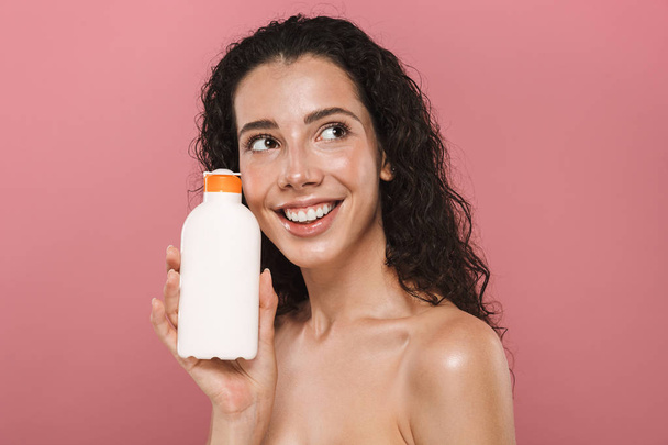 Portrait of pretty beautiful young woman posing isolated over pink background take care of her skin holding cream lotion gel. - Фото, изображение