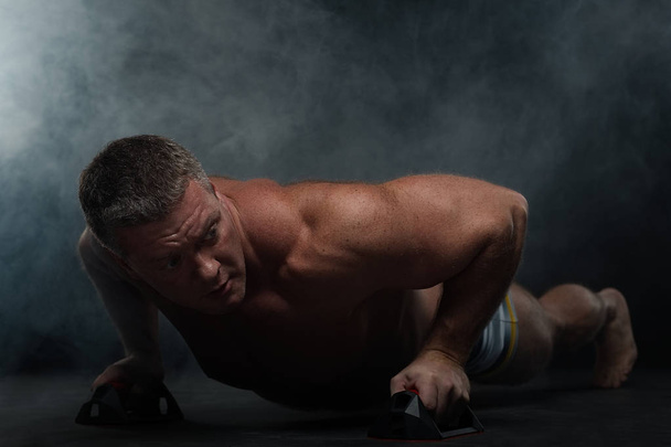 Powerful muscular man does pushups in the smoke on a black background - Фото, зображення