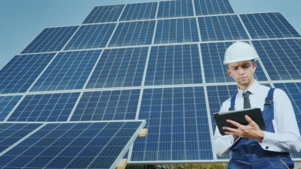 A young businessman in a white helmet uses a tablet on the background of solar panels - Filmati, video