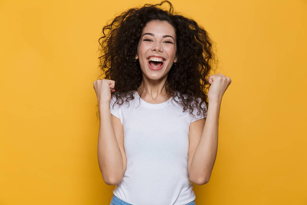 Image of young woman 20s with curly hair yelling and clenching fists isolated over yellow background - Photo, Image