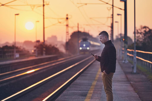 Young man waiting at railroad station platform and using smart phone against commuting train at sunrise. - Foto, afbeelding