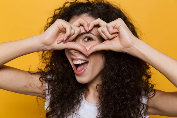 Photo of caucasian woman 20s with curly hair smiling and showing heart shape with fingers isolated over yellow background - Foto, Bild