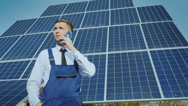 若いエンジニアは、大規模な太陽電池パネルの背景に電話で話す - 写真・画像