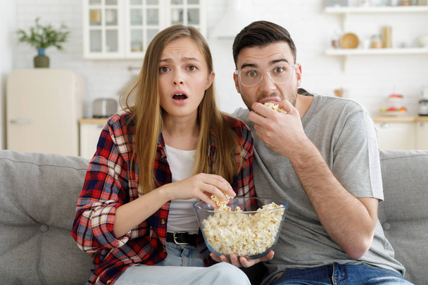 Surprised young couple watching TV sitting on couch at home with popcorn bowl  - Photo, Image