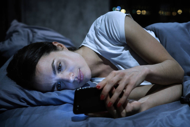 Smartphone addiction. Young tired female looking at her mobile phone screen, lying in bed late at night, scrolling her social media news feed - Photo, Image