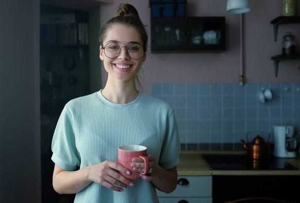 Late night shot of young pretty teen wearing round eyeglasses, holding pink cofee mug, looking straight to the camera, smiling with perfect white teeth - Фото, зображення