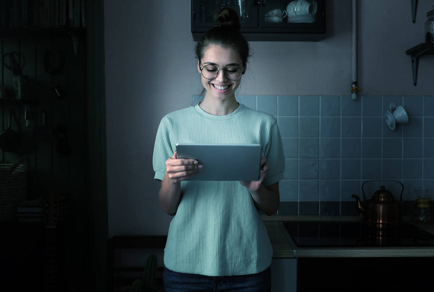 Young attractive smiling european female in kitchen with tablet in hands looking at screen through big trendy round eyeglasses at night - Zdjęcie, obraz