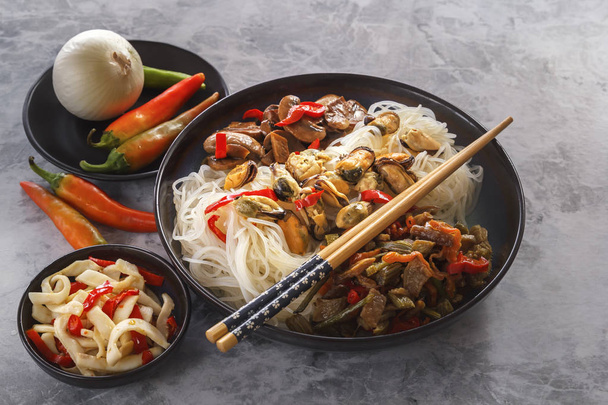 Rice noodles with seafood, salad, fried mushrooms and red pepper stand on a gray table. - Photo, Image