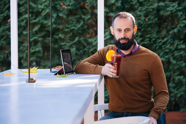 Un barbu dans un café buvant du vin chaud. Homme d'affaires travaille au bureau, tient un verre avec une boisson chaude rouge et utilise une tablette
. - Photo, image
