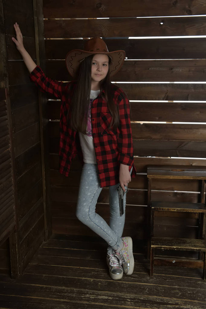 young girl cowboy in a red checkered shirt on the background of a wooden wall - Foto, Bild