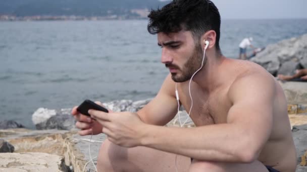 Young man at beach listening to music with earphones - 映像、動画
