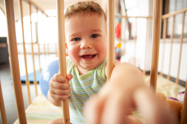 Bebê sorridente bonito olhando através das barras de madeira de seu berço ou playpen com um sorriso feliz dentro de casa no berçário
 - Foto, Imagem