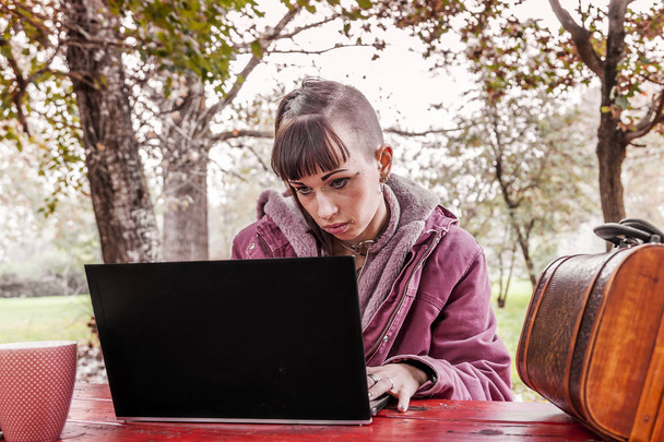 молода жінка навчається з комп'ютером, сидячи на дерев'яному столі в парку серед осіннього листя
 - Фото, зображення