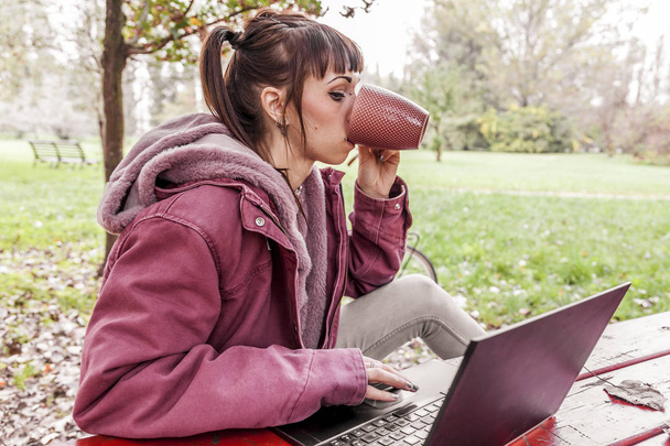 junge Frau, die am Computer lernt, sitzt auf einem Holztisch im Park inmitten der Herbstblätter - Foto, Bild