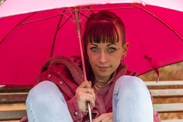 портрет молодої жінки, що ховається від осіннього дощу з червоною парасолькою, що сидить на лавці парку
 - Фото, зображення
