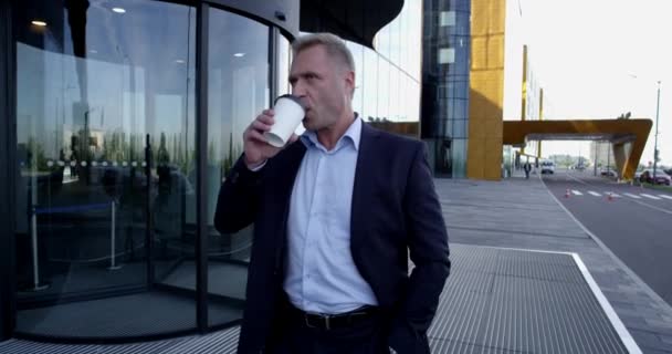 Mature business man walking outdoors along office building holding takeaway coffee - Filmagem, Vídeo