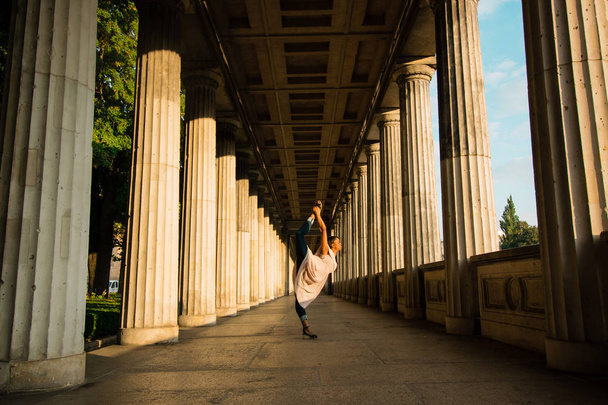 The young black beautiful woman with afro curly dark hair making yoga pose in the cityscape, variation of Natarajasana. Woman wearing gentle beige dress and dancing near big colums in the sunrise.  - Photo, Image