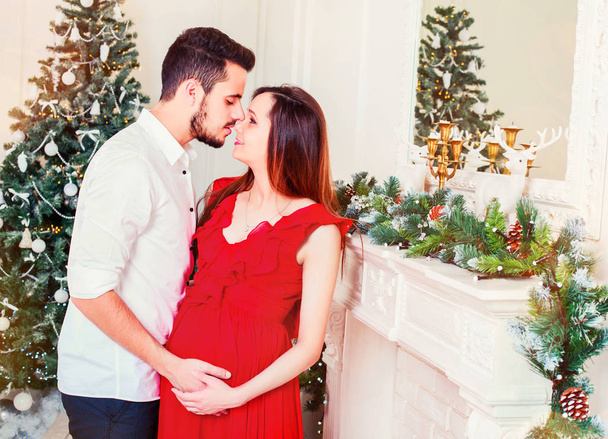 Happy family couple on Christmas at fireplace. Happy couple kiss and hugging. Living room decorated by Xmas tree and present gift box, the light give cozy atmosphere. Waiting for baby. New Year theme. Toning - Photo, Image