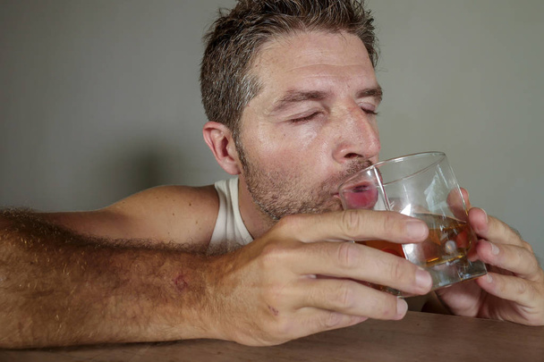 young drunk and pissed alcoholic man wasted wearing dirty singlet drinking whiskey glass intoxicated and messy isolated on dark background in alcohol abuse and addiction and alcoholism problem - Foto, Bild