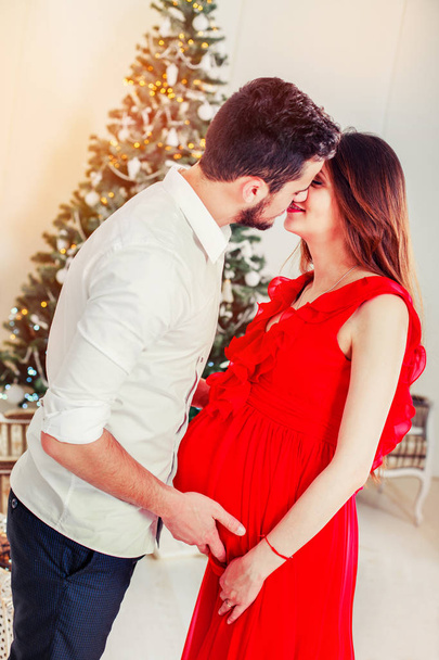 Happy family couple on Christmas at fireplace. Happy couple kiss and hugging. Living room decorated by Xmas tree and present gift box, the light give cozy atmosphere. Waiting for baby. New Year theme - Фото, зображення