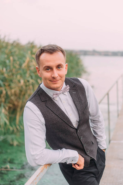 Young happy relaxed fair-haired man with blue eyes looks at the camera and smiles, stands on the bridge near a small river. Portrait of a man, groom in a gray wedding suit. art photo. - Foto, Bild