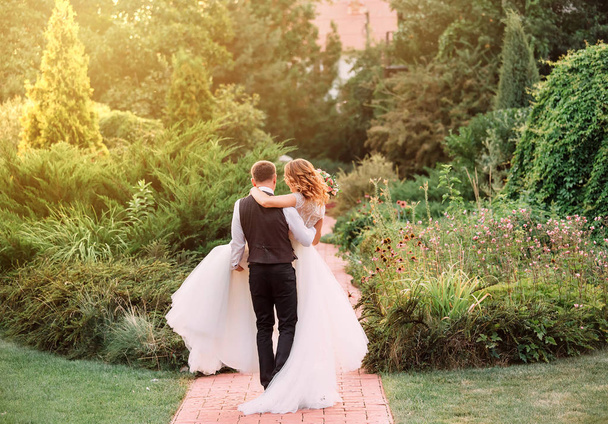 young bridegroom in a suit carries in his arms his bride, dressed in a long magnificent luxurious white dress, in an amazing garden. no faces in the picture, beautiful romantic photo, art processing. - Photo, Image