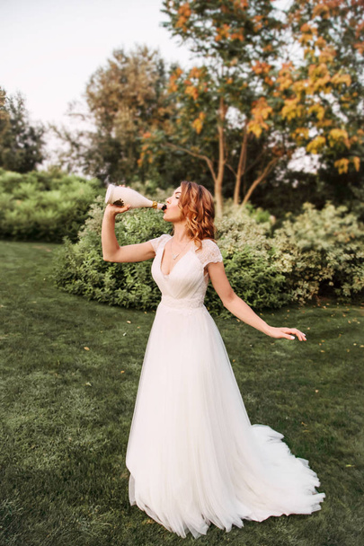 young redhead girl in a white elegant wedding long dress drinking champagne from a beautifully decorated bottle - Фото, зображення
