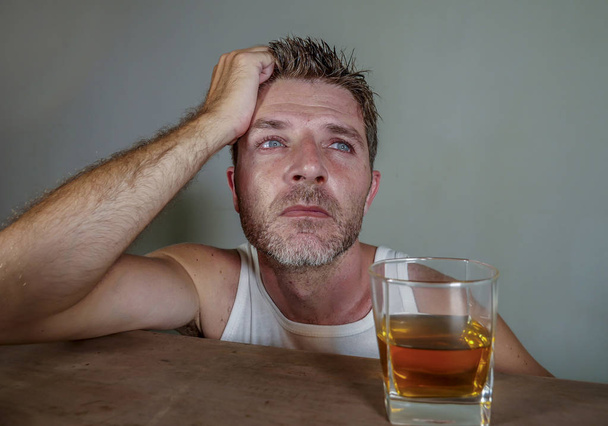 young wasted and depressed alcohol addict man in dirty singlet drinking glass of whiskey feeling desperate suffering alcoholism problem and booze addiction on isolated background in alcoholic concept - Фото, изображение