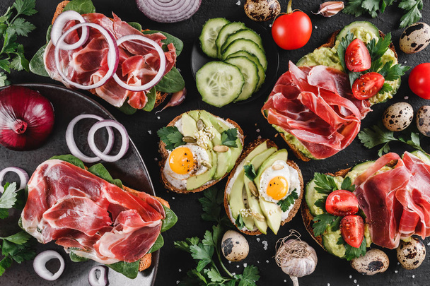 Various of sandwiches and bruschetta with prosciutto, fried quail egg, avocado, cucumber, tomatoes, spices and greens on black stone background. Clean eating, healthy breakfast. Top view, flat lay - Photo, Image