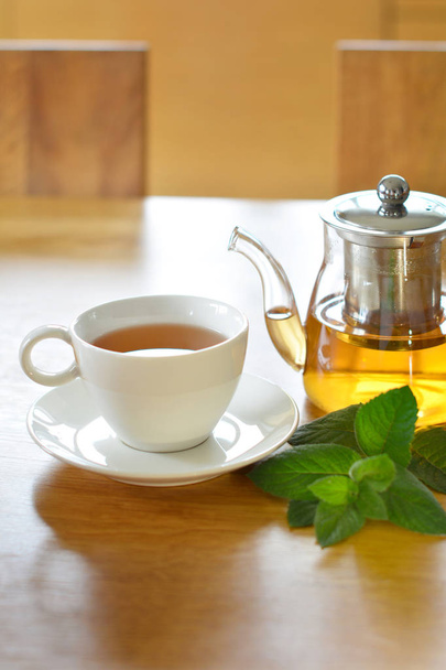 Белая керамическая чашка и стеклянный чайник травяного чая со свежей веточкой мяты на деревянном столе
 - Фото, изображение