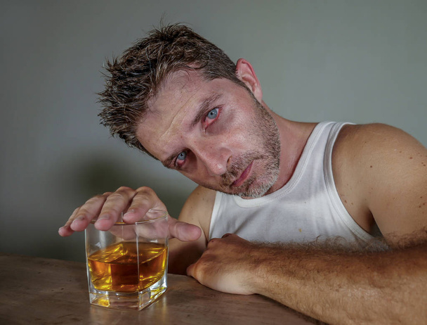 young drunk and pissed alcoholic man wasted wearing dirty singlet drinking whiskey glass intoxicated and messy isolated on dark background in alcohol abuse and addiction and alcoholism problem - Photo, image