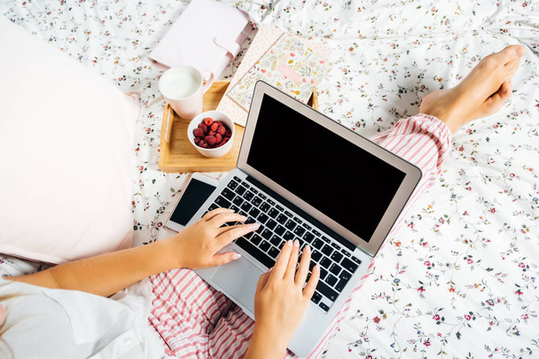 Young woman drinking milk at home in bed and checking her laptop, top view - Foto, Imagem