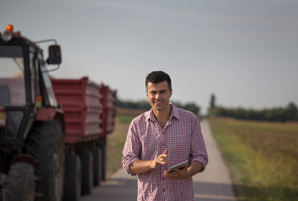 Beau fermier avec tablette debout devant le tracteur avec remorques dans le champ
 - Photo, image