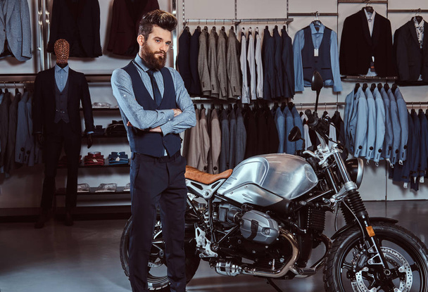 Elegancko ubrany mężczyzna pozowanie z skrzyżowanymi rękami w pobliżu retro sportowy motocykl sklep Odzież męska. - Zdjęcie, obraz