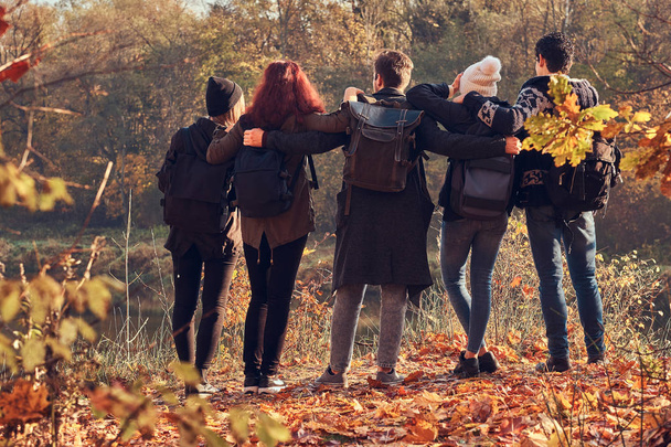 Back view of young friends hugging together and looking at the lake in beautiful autumn forest. - Photo, Image