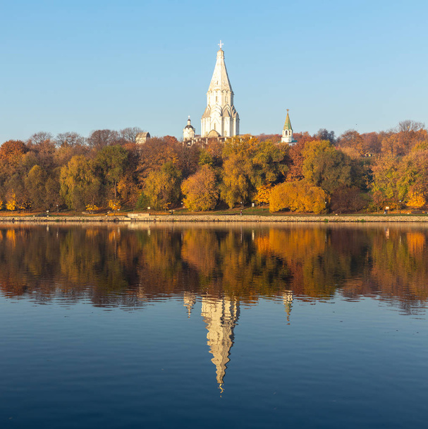 Colorful autumn. Alley along the riverbank on the background of the autumn park. Reflections in the water of the forest and blue sky. Over all towers the Orthodox Church of the Ascension - Fotografie, Obrázek