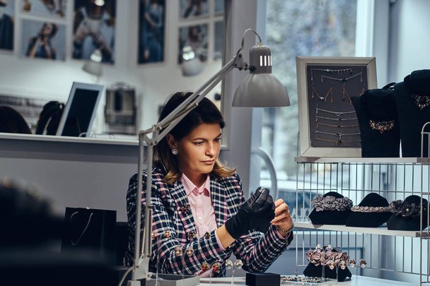 Trabalhadora de jóias feminina elegantemente vestida sentada à mesa de trabalho e fazer exame de jóias
. - Foto, Imagem