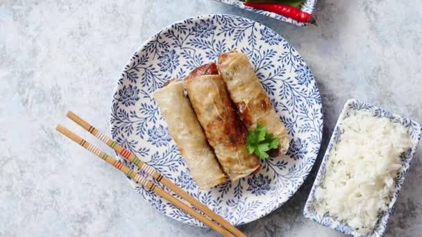 Orginal asian deep fried spring rolls placed on beautiful oriental style plate with chopstick, boiled rice, cucomber nad hot chilli peppers od side. Top view on stone background. - Footage, Video